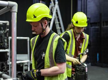 Two production & technical arts students in hard hats and high vis jackets organising lights in the Sainsbury Theatre at LAMDA