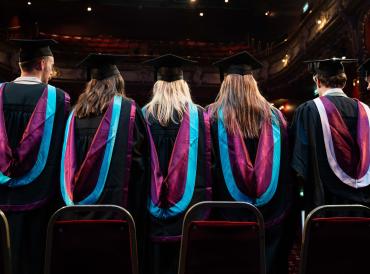 Photo of the backs of five students in academic gowns at the Lyric Hammersmith for the LAMDA 2023 graduation ceremony