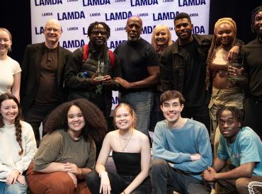 LAMDA students, Professor Mark O'Thomas and poet Roy McFarlane in the Sainsbury Theatre, smiling for a group photo at LAMDA's 2023 Poetry Night.,