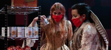 Two women in period dress wearing face masks shop for food tins