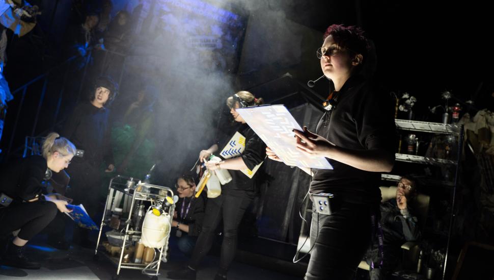 A student wearing a headset with mic and holding a clipboard on a busy stage