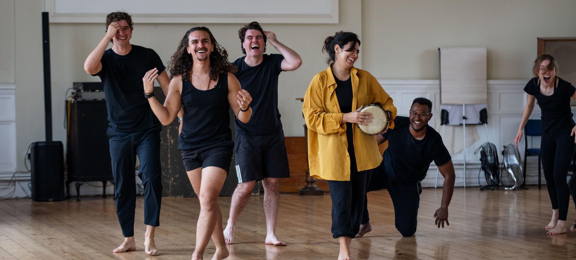 A group of performers laughing and dancing barefoot in a rehearsal studio, one playing a hand drum