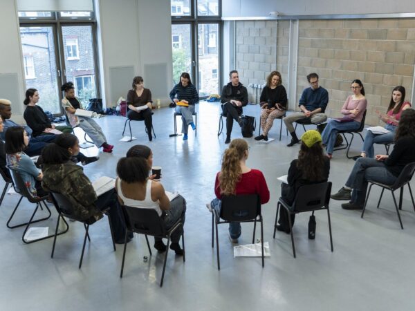 A group sits in a circle of chairs holding scripts, taking part in a rehearsal or workshop discussion in a bright studio space