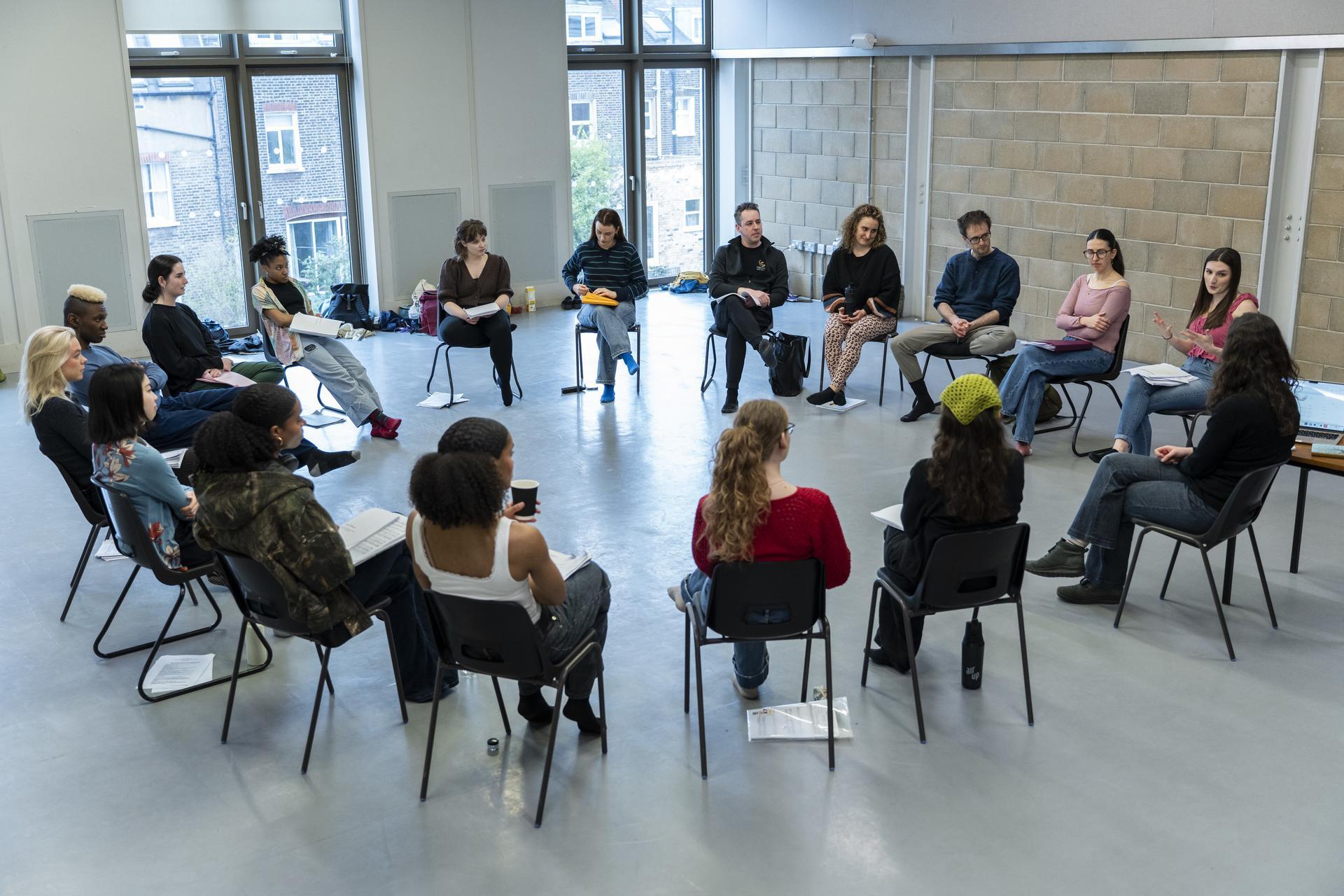A group sits in a circle of chairs holding scripts, taking part in a rehearsal or workshop discussion in a bright studio space