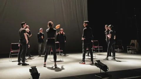 Performers dressed in black stand in small groups on a stage, rehearsing under spotlights, with empty chairs nearby