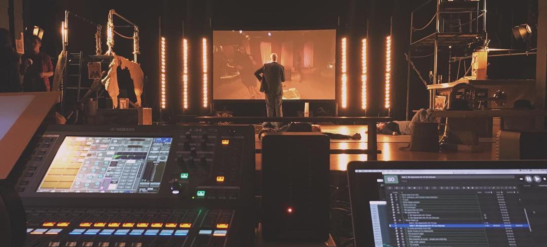 Sound and lighting control desk facing a stage with performers and a large screen backdrop