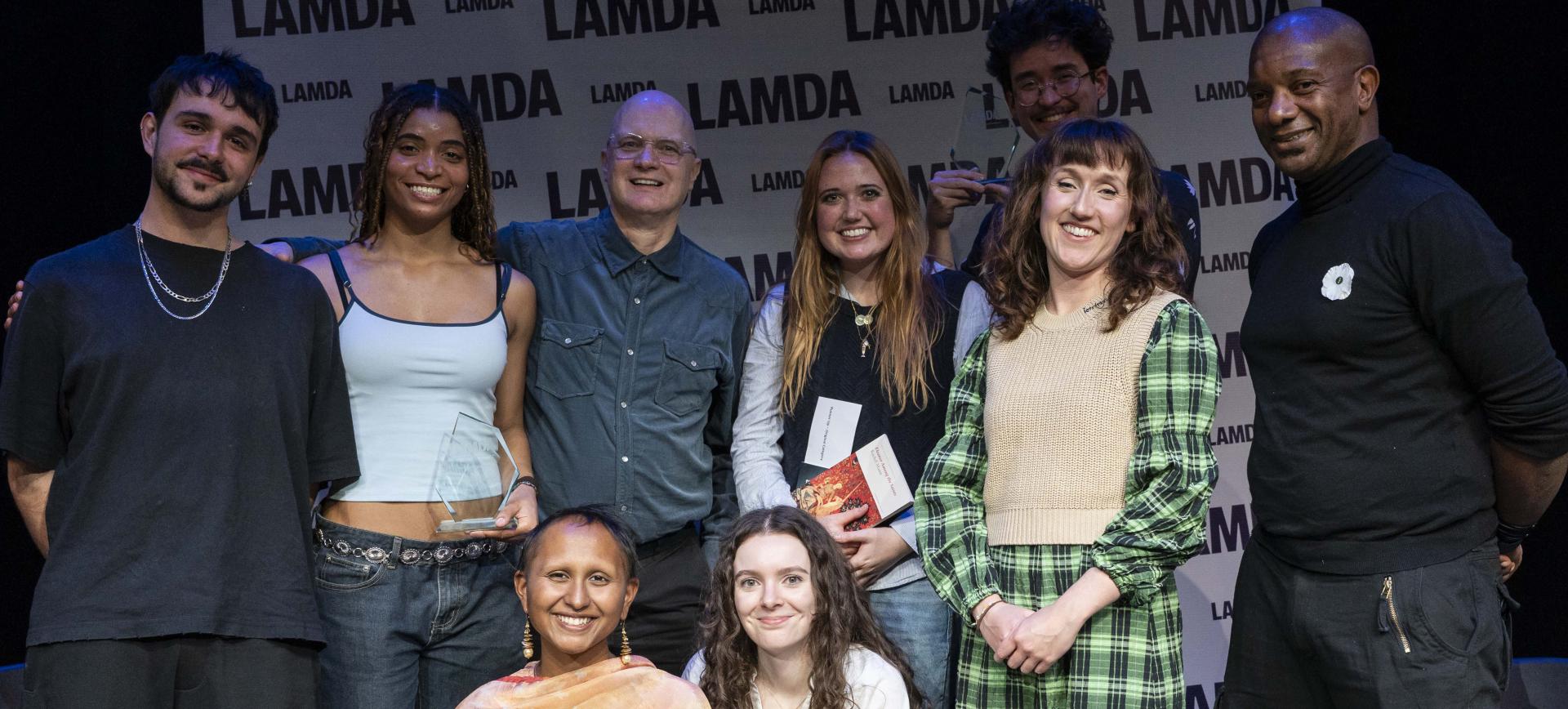 Group of award winners and presenters smiling together on stage in front of a LAMDA branded backdrop, one holding a glass trophy