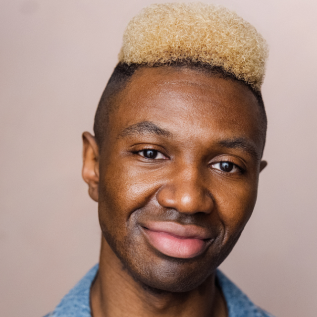 Close-up portrait of a smiling man with bleached blond curly hair, wearing a denim shirt