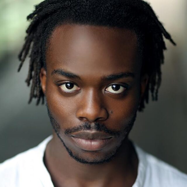 Close-up portrait of a young man with locs and a moustache, wearing a white top, looking directly at the camera