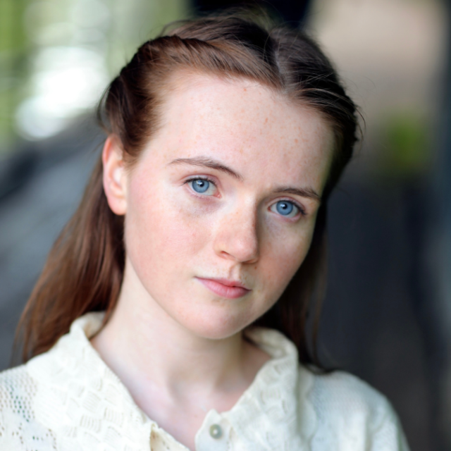 Close-up portrait of a young woman with red hair and blue eyes, wearing a cream knitted cardigan