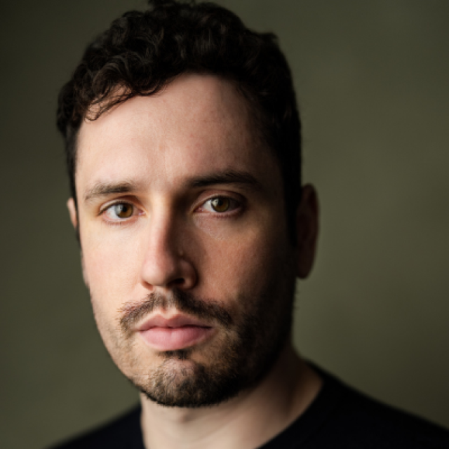 Close-up portrait of a man with curly dark hair and a beard, looking directly at the camera