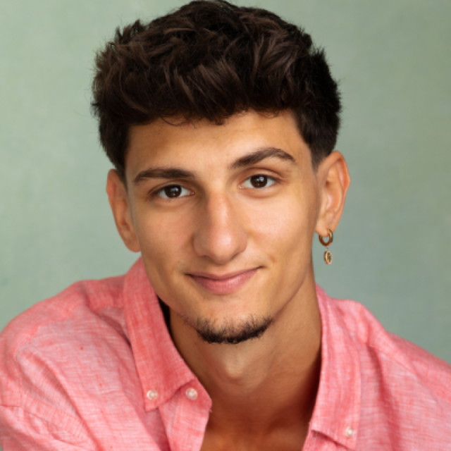 Portrait of a young man with curly dark hair and earrings, wearing a pink shirt, smiling at the camera
