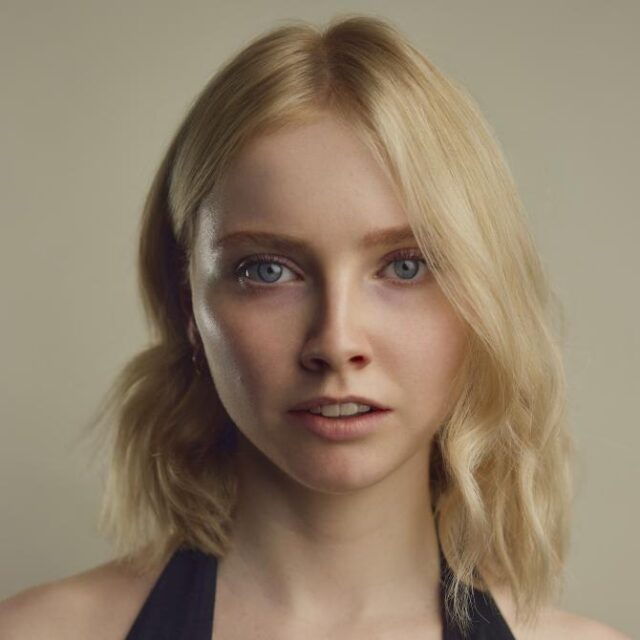 Close-up portrait of a young woman with blonde wavy hair and blue eyes, looking directly at the camera