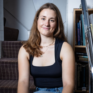 Young woman with long brown hair smiling, sitting near a staircase and bookshelf