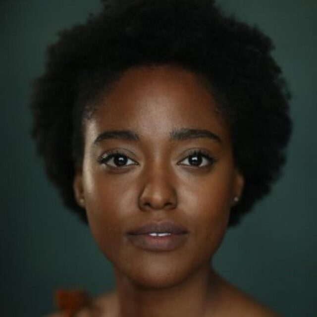 Close-up portrait of a woman with natural curly hair, looking directly at the camera against a dark green background