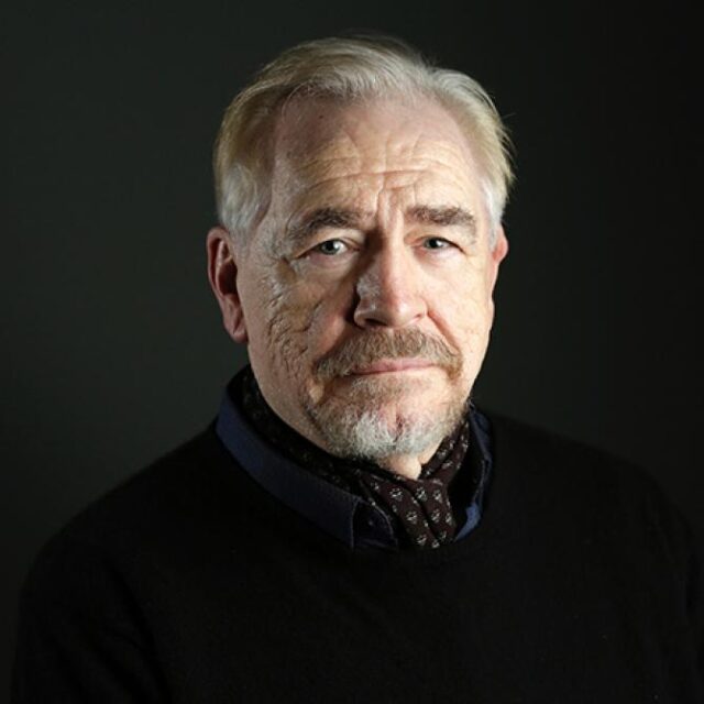 Portrait of an older man with grey hair and a moustache, wearing a dark jumper against a dark background