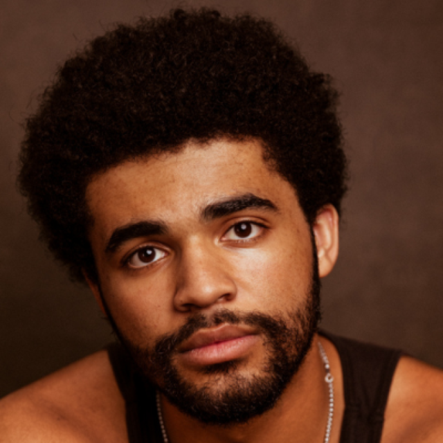 Close-up portrait of a young man with a curly afro and beard, wearing a chain necklace