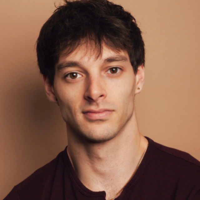 Portrait of a young man with short dark hair, wearing a maroon T-shirt and a thin gold chain necklace
