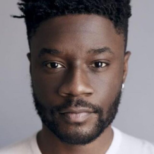 Close-up portrait of a man with short curly hair and a beard, wearing a white top, against a plain grey background