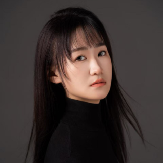 Studio portrait of a young woman with long dark hair and fringe, wearing a black turtleneck, against a grey background