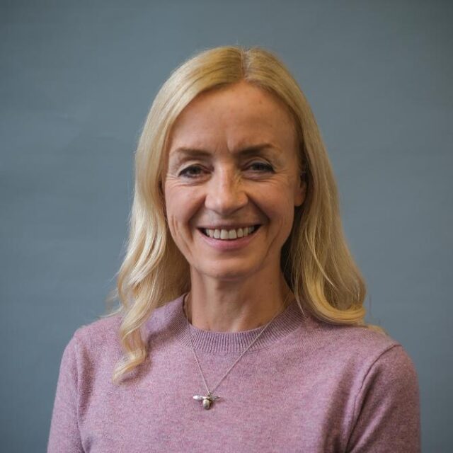 Portrait of a smiling blonde woman wearing a pink jumper and a bee-shaped pendant necklace