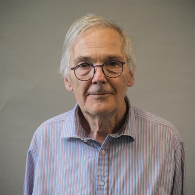 Older man with grey hair and glasses, wearing a striped shirt, against a plain background