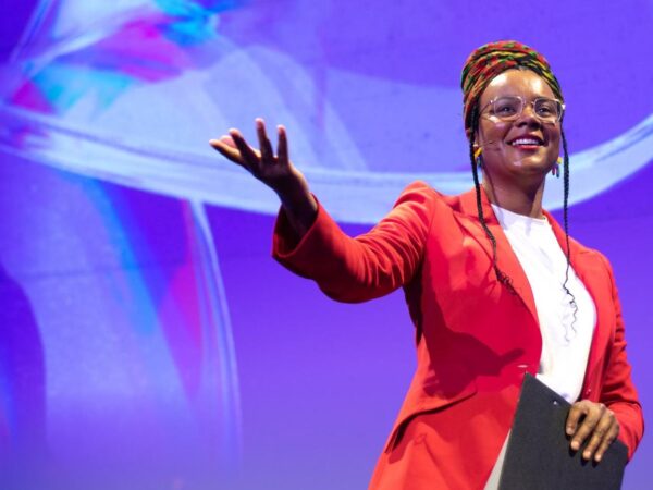 Speaker in a red blazer and headwrap gestures while presenting on stage against a purple backdrop
