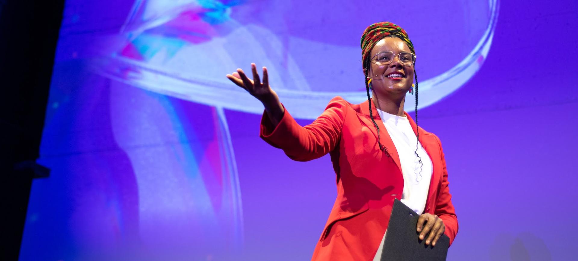 Speaker in a red blazer and headwrap gestures while presenting on stage against a purple backdrop