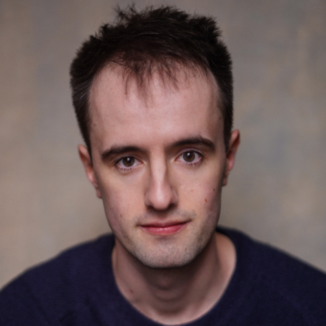 Close-up portrait of a young man with short dark hair, wearing a dark jumper, against a mottled grey background