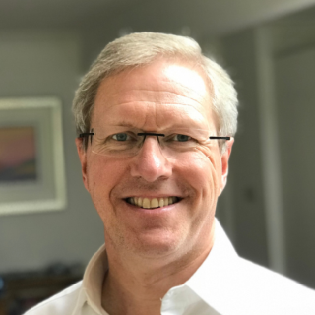 Older man with grey hair and glasses, smiling at the camera, wearing a white shirt