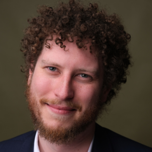 Portrait of a smiling man with curly brown hair and a beard, wearing a dark jacket