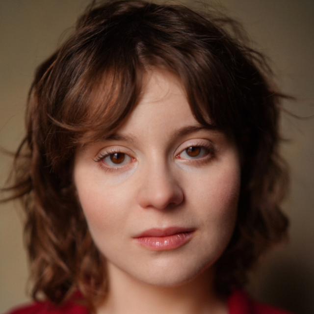 Close-up portrait of a woman with curly brown hair, looking directly at the camera with a neutral expression
