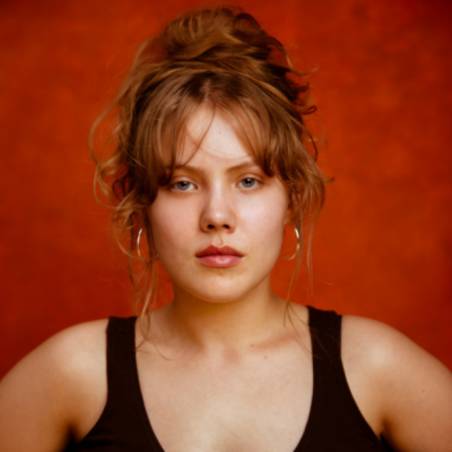Portrait of a young woman with red hair in an updo and hoop earrings, against a deep orange background