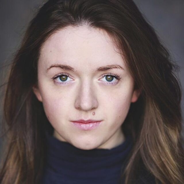 Close-up portrait of a young woman with long brown hair, looking directly at the camera with a neutral expression