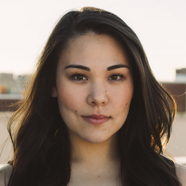 Close-up portrait of a young woman with long dark hair, outdoors against a bright sky