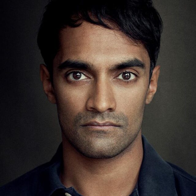 Close-up portrait of a man with dark hair and stubble, looking directly at the camera with a serious expression