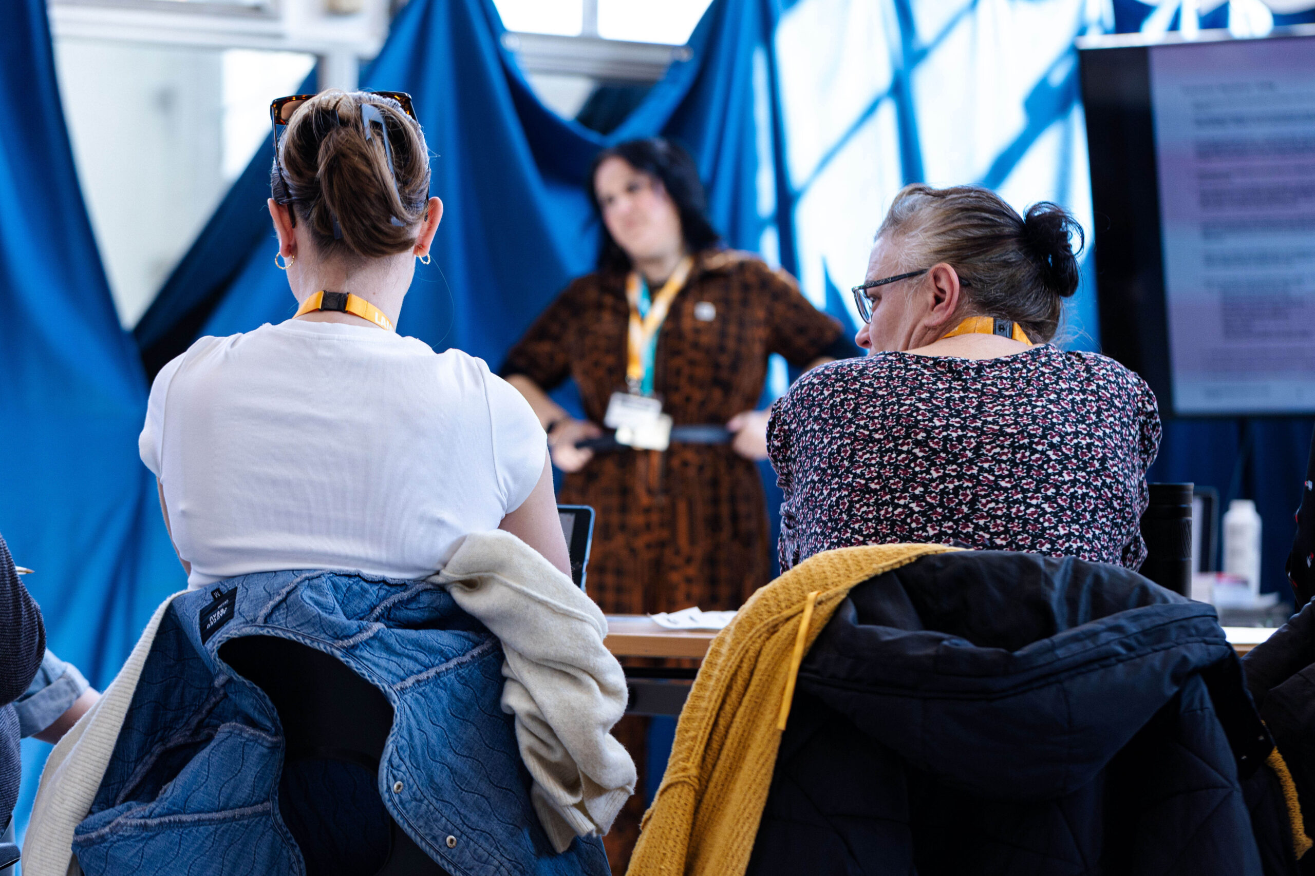 Attendees wearing conference lanyards sit facing a presenter adjusting her lanyard at a poster session with blue drapes