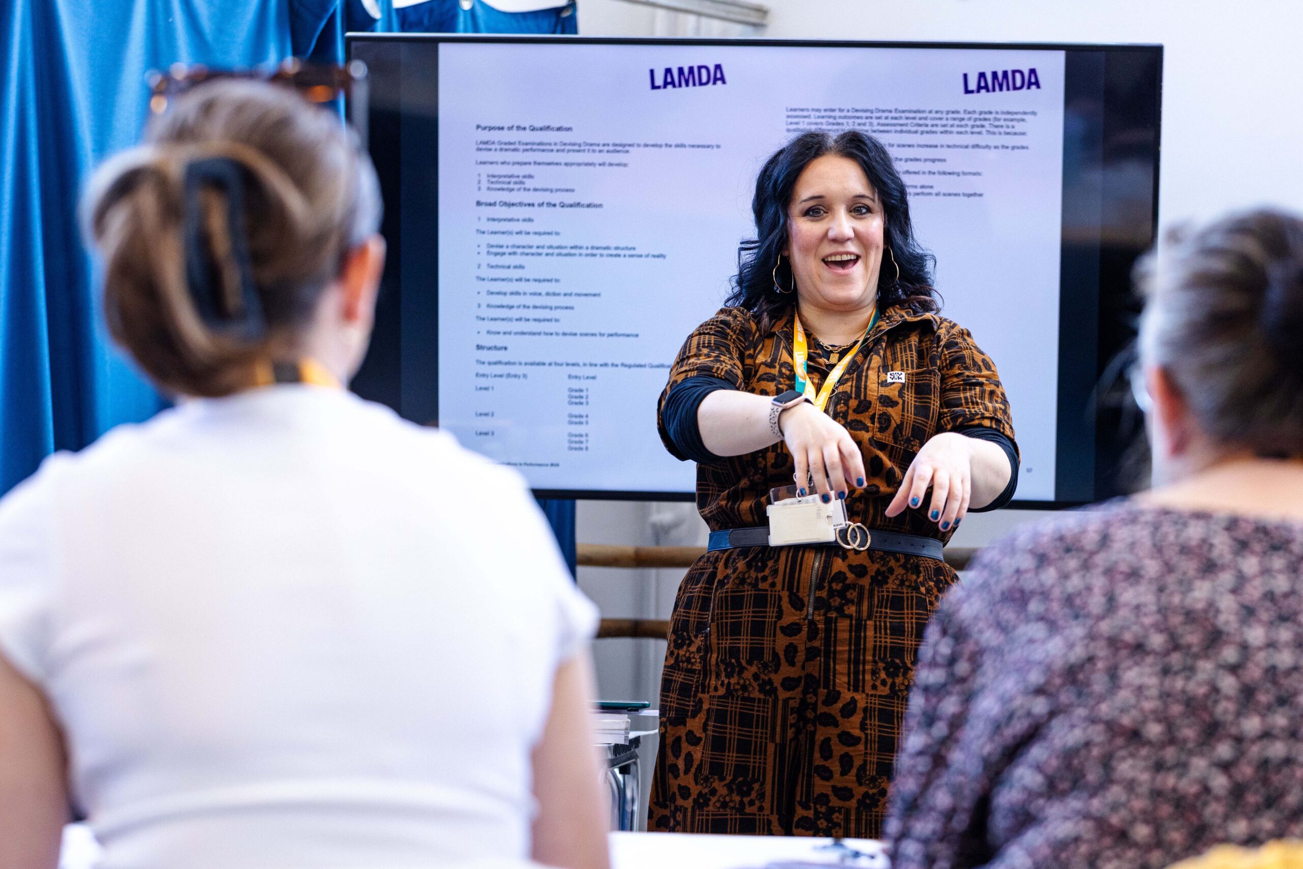 A trainer gestures while presenting to an audience, with a screen behind her showing LAMDA Devising Drama qualification details