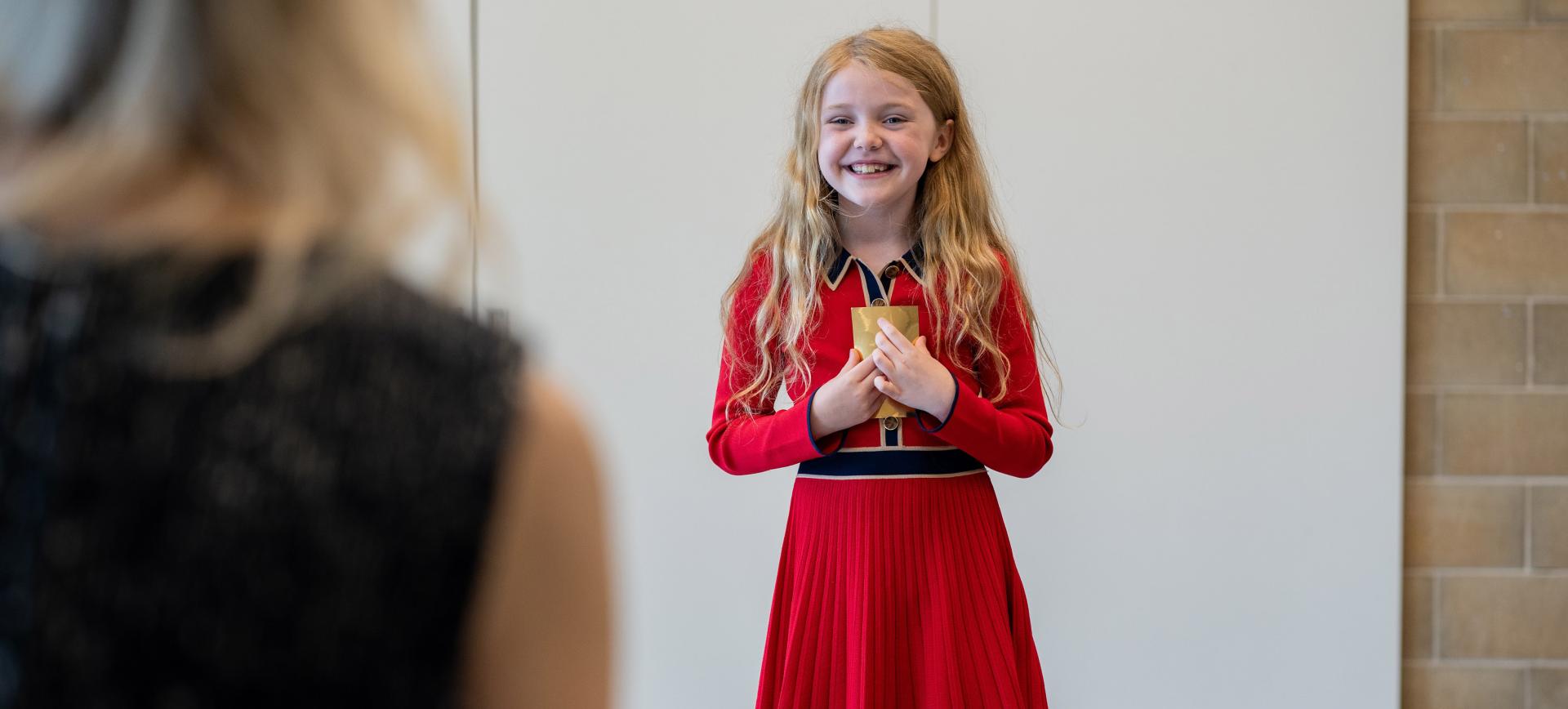 Young girl in a red dress smiling and holding a small gold gift box to her chest
