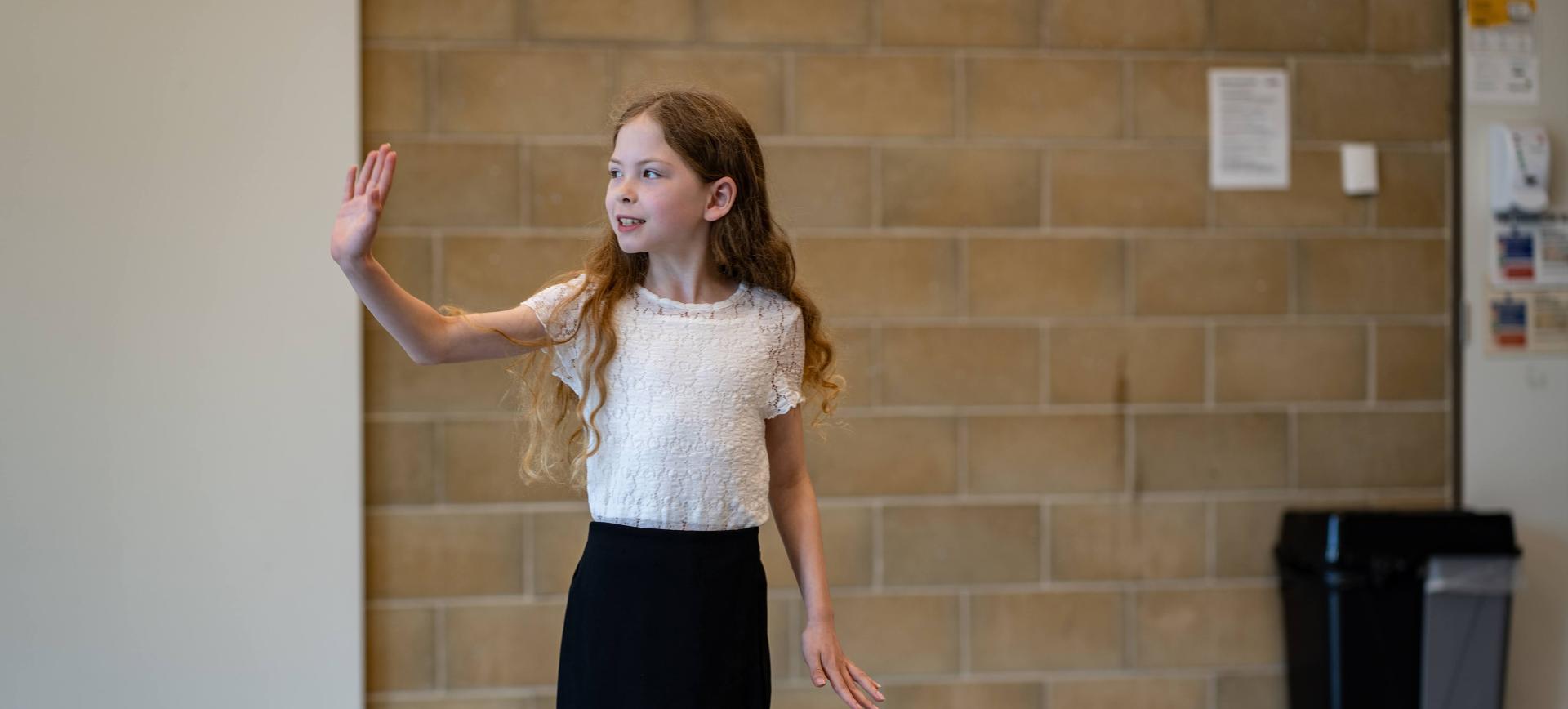 Girl with long hair raises her hand while speaking, standing in a corridor