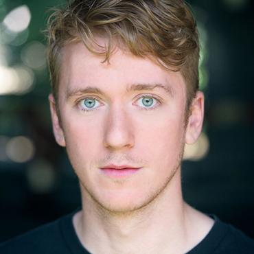 Headshot of a young man with light brown hair and blue eyes, wearing a black top, against a blurred outdoor background