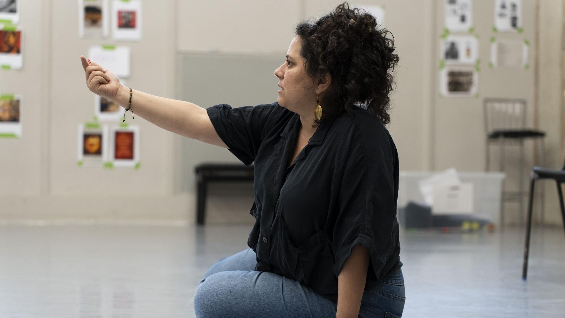 Woman sitting cross-legged on a studio floor, gesturing upward while directing a rehearsal, with a script folder beside her