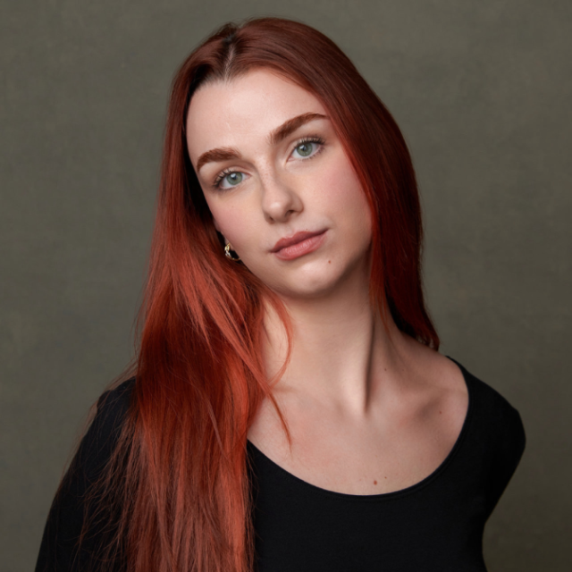 Woman with long red hair and a black top, looking directly at the camera against a plain grey background
