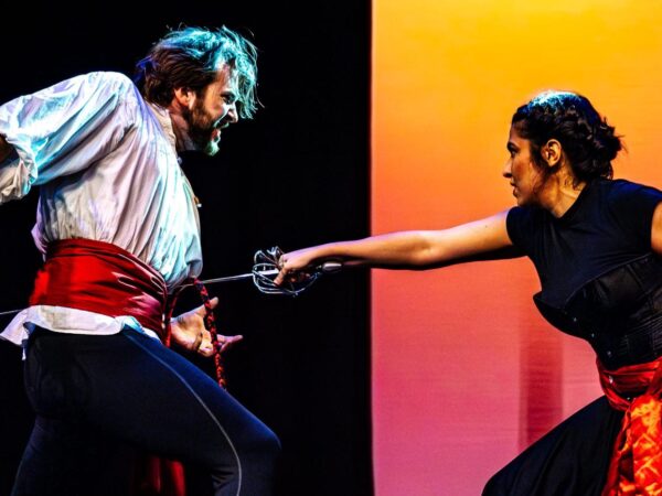 Two performers duel with swords on stage, blades crossed, faces intense against a dramatic orange-lit backdrop