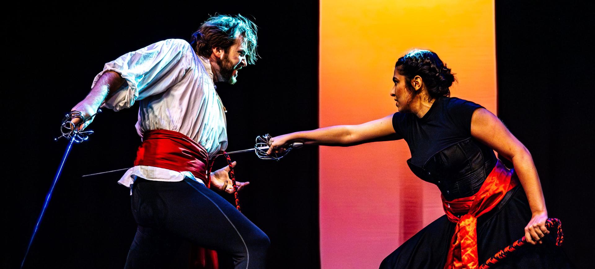 Two performers duel with swords on stage, blades crossed, faces intense against a dramatic orange-lit backdrop