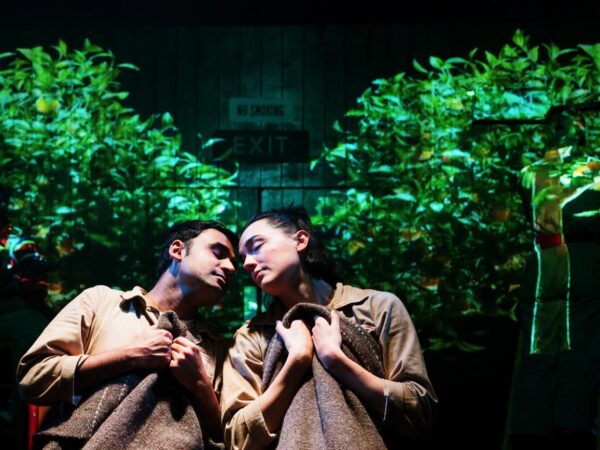 Two actors in beige costumes stand head to head clutching blankets, lit against a projected green foliage backdrop on stage