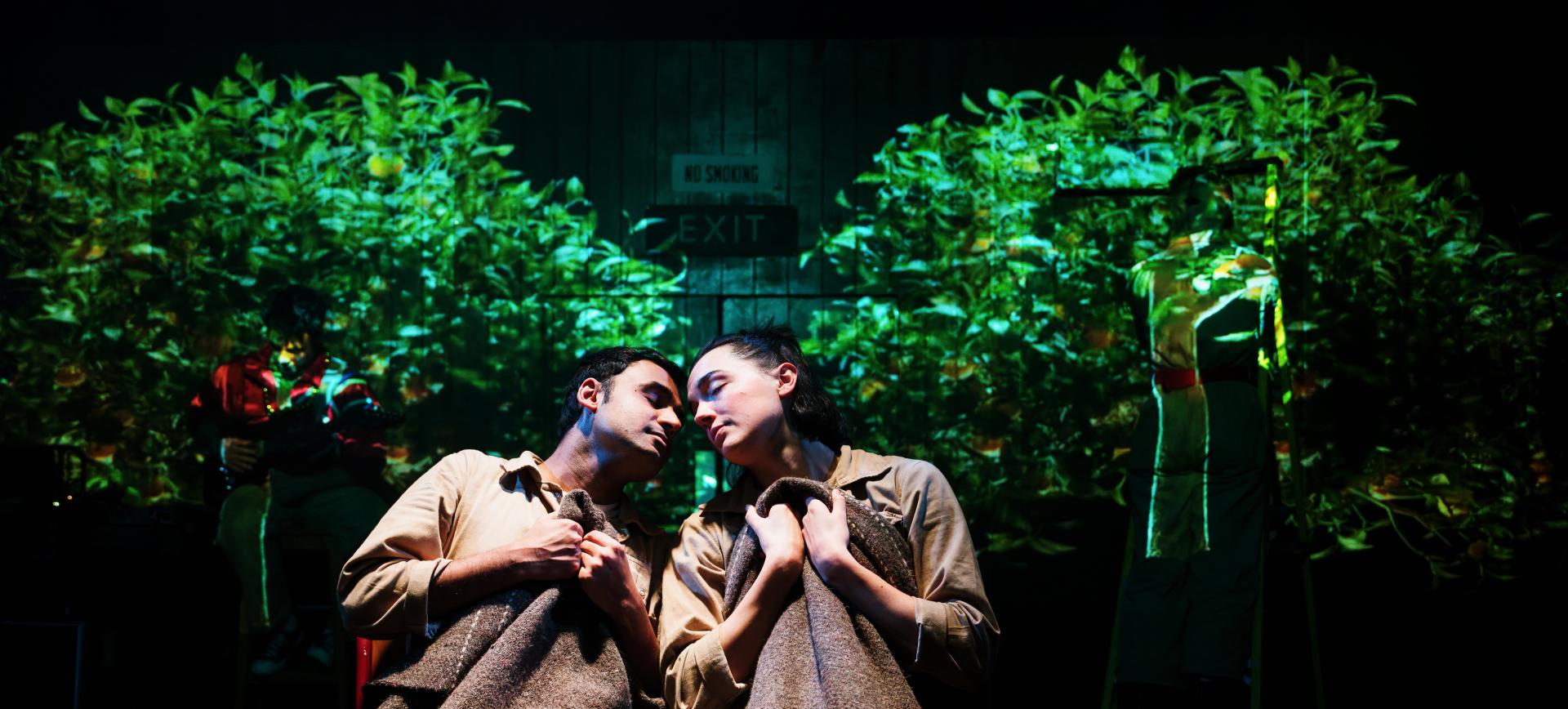 Two actors in beige costumes stand head to head clutching blankets, lit against a projected green foliage backdrop on stage