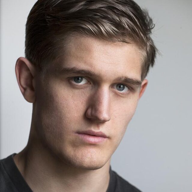 Close-up portrait of a young man with light brown hair and blue eyes, looking directly at the camera with a neutral expression