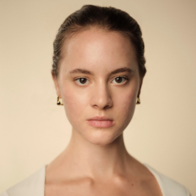 Close-up portrait of a young woman with slicked-back hair, gold earrings and a neutral expression, facing the camera directly