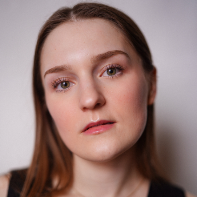 Close-up portrait of a young woman with fair skin and long brown hair, looking directly at the camera with a neutral expression